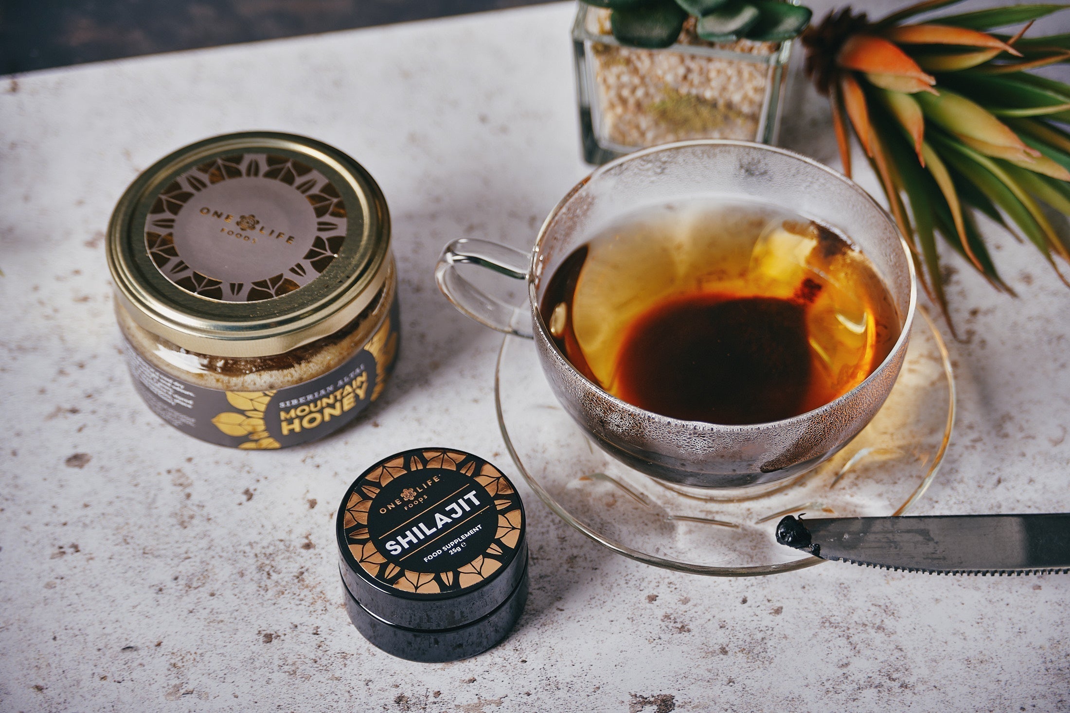 Glass cup of dark tea beside jars of One Life Foods Shilajit resin and clover honey, with a knife holding a small amount of Shilajit on a stone surface.