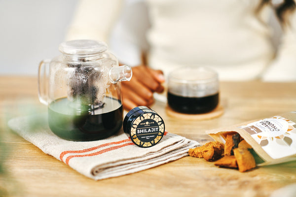 One Life Foods Shilajit resin and Chaga mushroom pieces beside a teapot and cup on a wooden table.