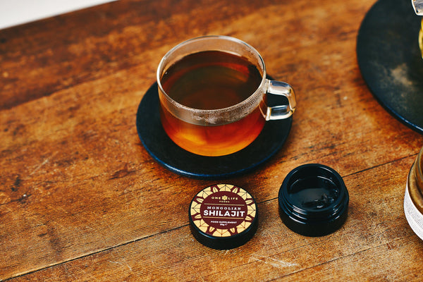 One Life Foods Shilajit resin jar shown next to a freshly brewed cup of tea.