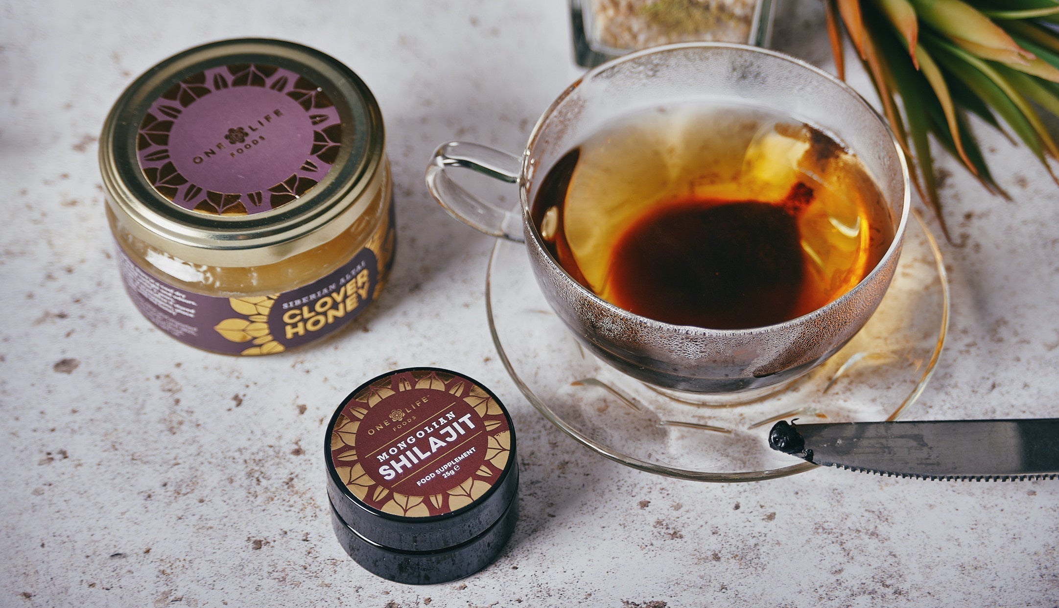 One Life Foods Shilajit resin and clover honey beside a glass cup of green tea.