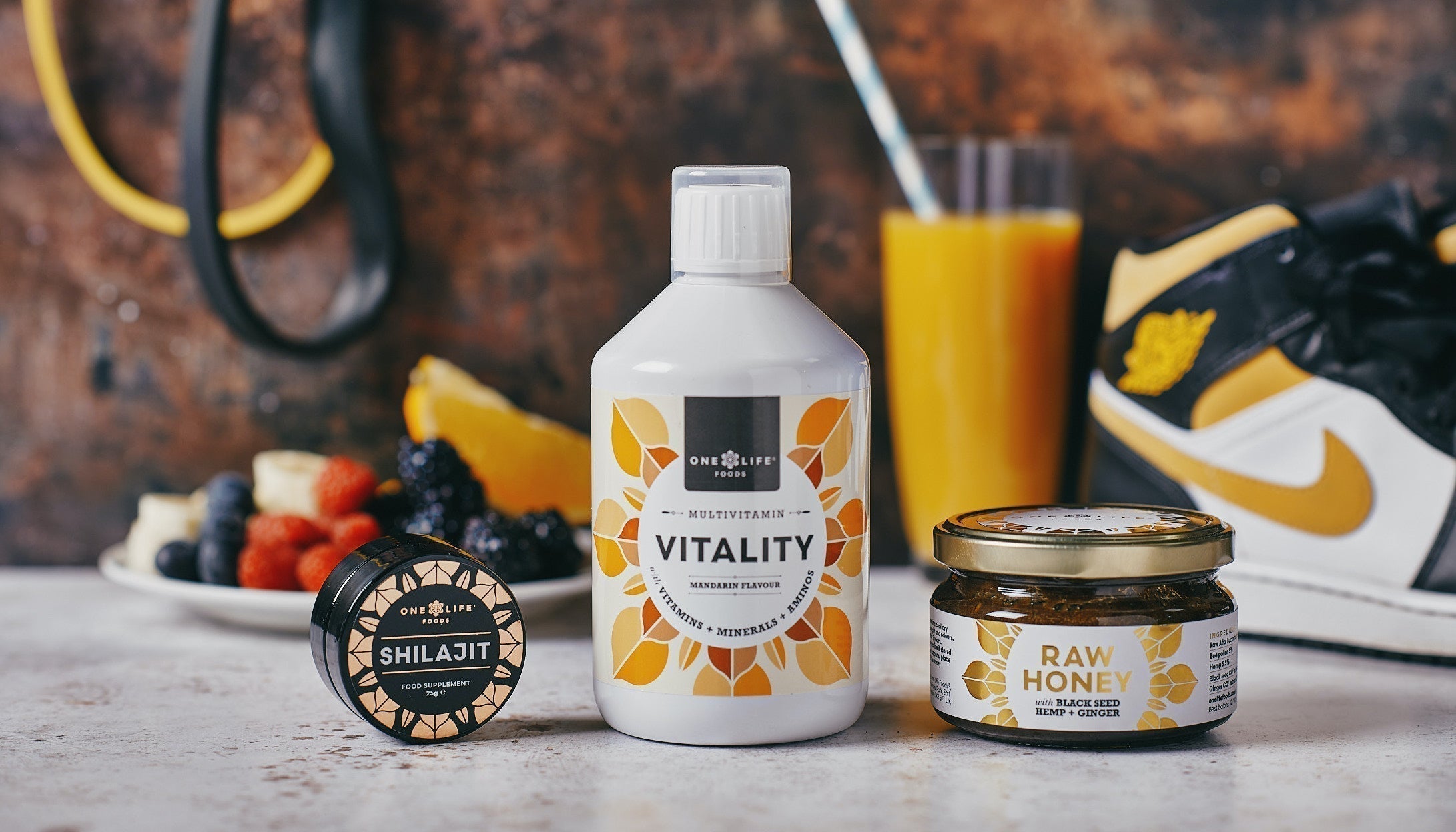 One Life Foods Vitality, Raw Honey, and Shilajit products displayed with fresh fruit, orange juice, and trainers in the background, representing active daily nutrition.