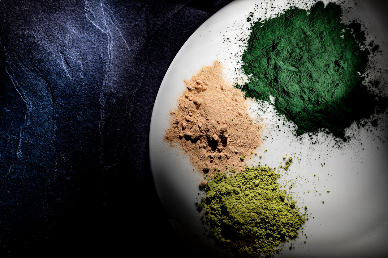 Assorted natural herbal supplement powders in shades of green and brown on a white plate against a dark background