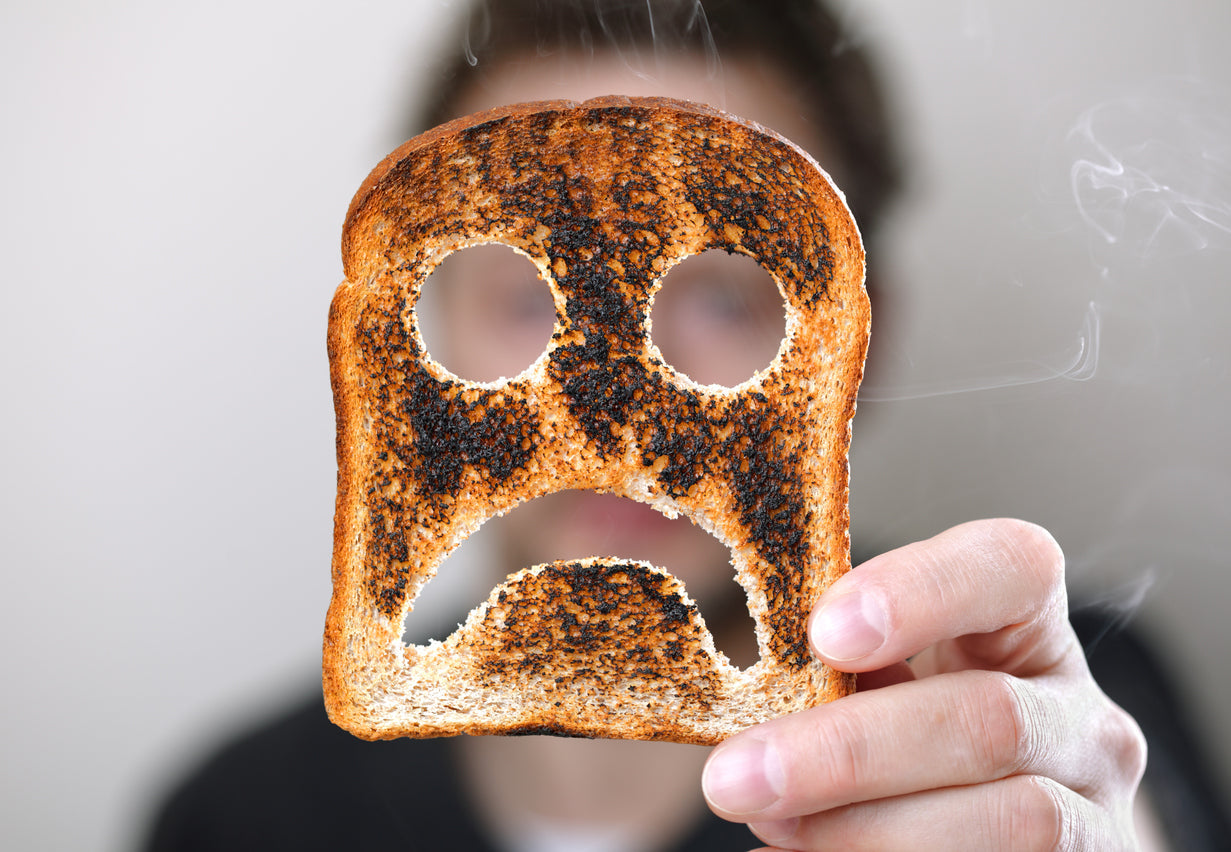 Person holding a burnt slice of toast with cut-out eyes and a frowning mouth, resembling a sad face, against a blurred background.