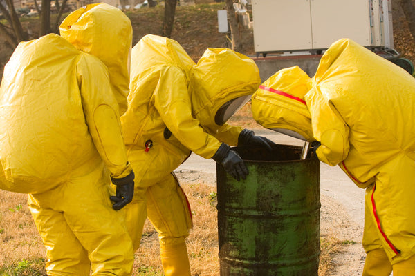 Team of people wearing yellow hazmat suits inspecting a green barrel outdoors.