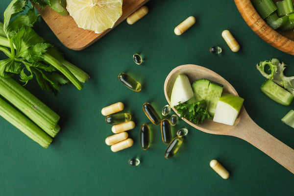Assorted supplements and green vegetables on a dark green background with a wooden spoon of fresh cucumber and herbs.