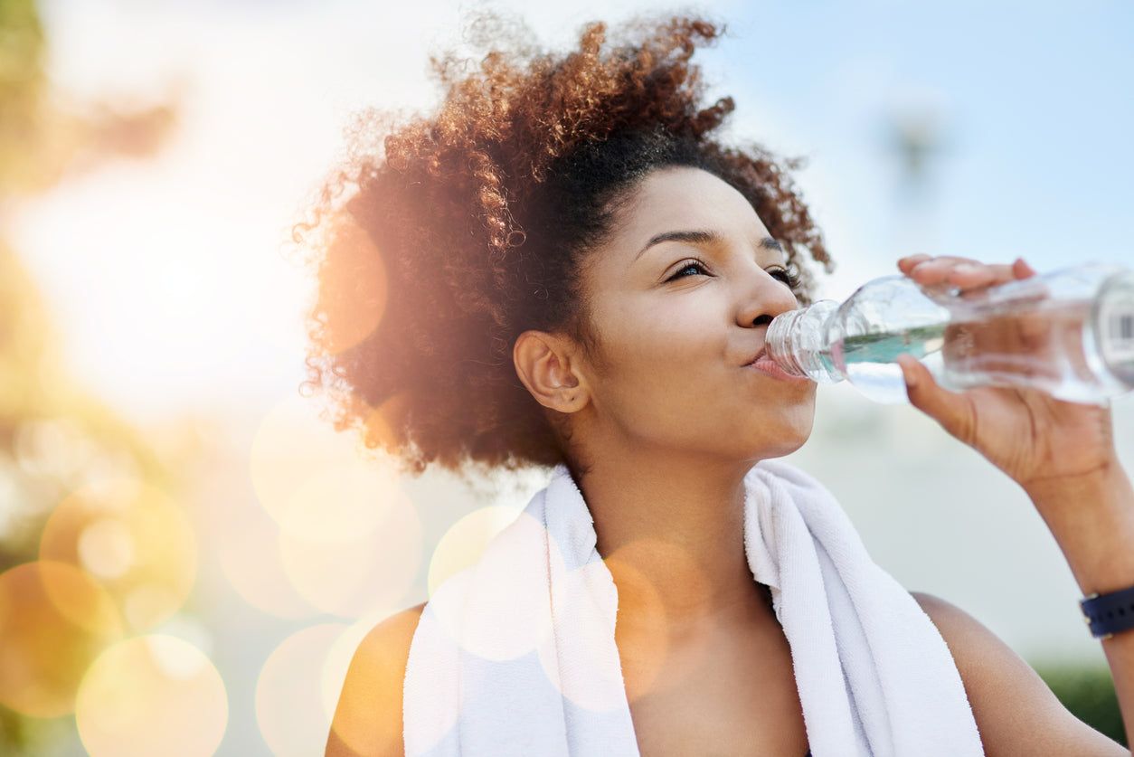 Woman rehydrating after exercise, drinking water outdoors in sunlight.