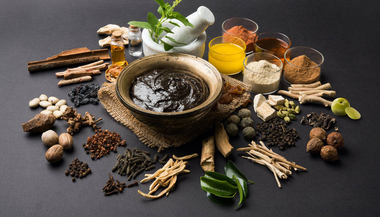 Bowl of herbal paste with assorted Ayurvedic herbs, roots, and powders on a dark background.