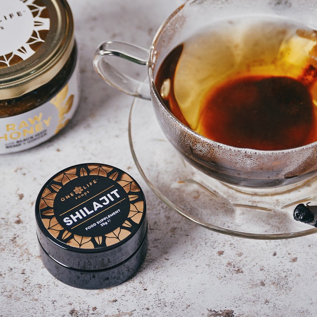 One Life Foods shilajit resin beside a glass of tea and raw honey.